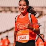 Smiling girl running in a marathon, giving a thumbs up, wearing a bright orange shirt.